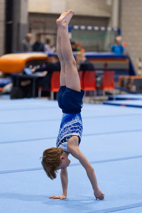 Young gymnast performs a vertical handstand on floor exercise mat, legs straight and pointed upward in blue patterned leotard