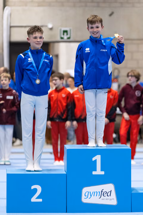 Two young gymnasts in blue tracksuits stand on medal podium, first and second place winners displaying their medals with pride