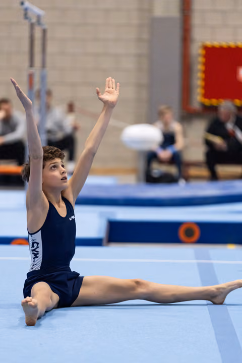 Young gymnast in navy leotard performs floor routine with extended splits pose, arms raised gracefully overhead in gymnasium