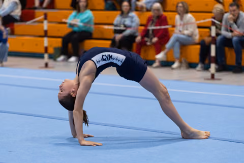 Gymnast performs a dramatic backbend on blue mat while spectators watch from bleachers in gymnasium
