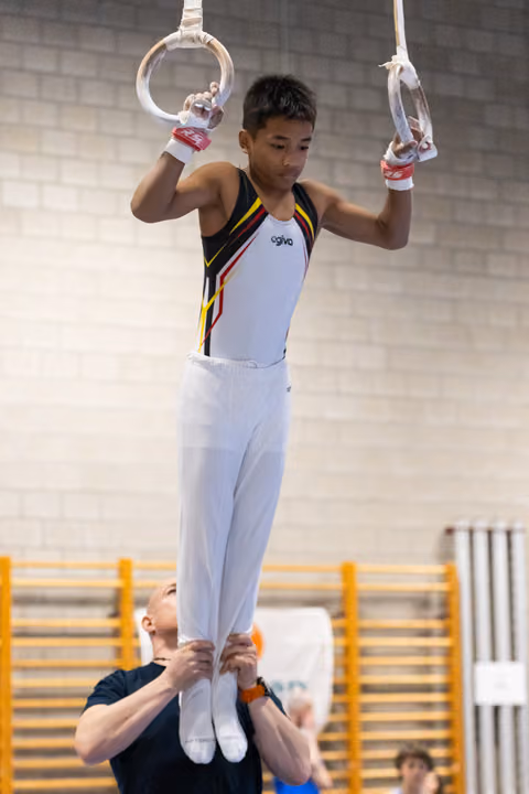 Young gymnast performs rings routine while coach spots from below in training hall