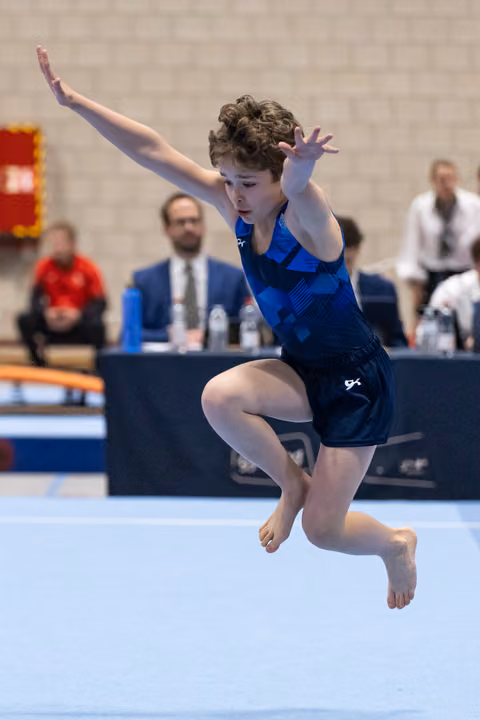 Young gymnast performs an expressive floor routine, suspended mid-air with arms raised and knees bent during her tumbling pass.