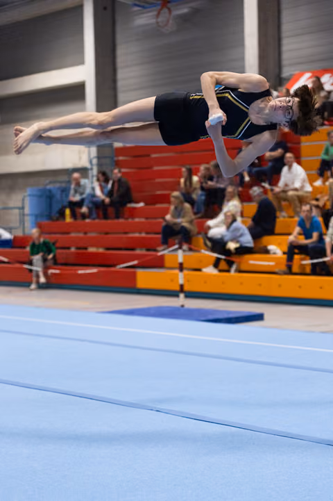 Gymnast performs horizontal backflip above blue floor mat with spectators watching from colorful bleachers