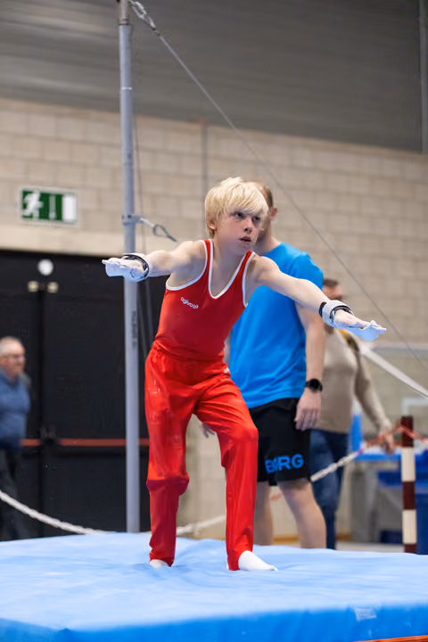 Young gymnast in red uniform dismounts from parallel bars with intense focus, coach observing in background