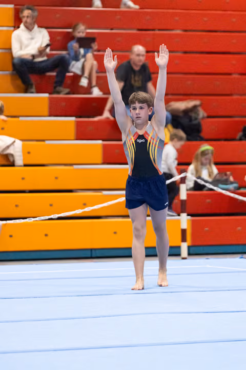 Young gymnast in navy leotard stands with arms raised in salute position on blue floor mat, spectators in bleachers behind