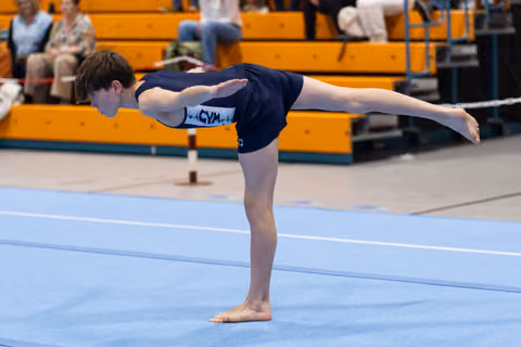 Young gymnast performs a horizontal balance pose on the floor exercise mat with spectators watching from the bleachers