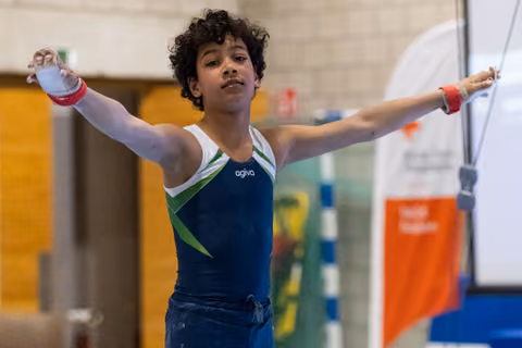 Young gymnast performs on rings with arms extended, showing concentration and control in blue and green leotard