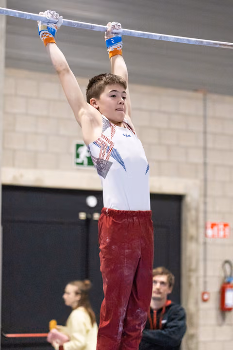 Young gymnast grips the high bar with focused expression, wearing blue wrist guards and red pants during training