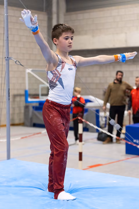 Young gymnast in white and burgundy uniform strikes an artistic pose with arms extended on blue competition mat