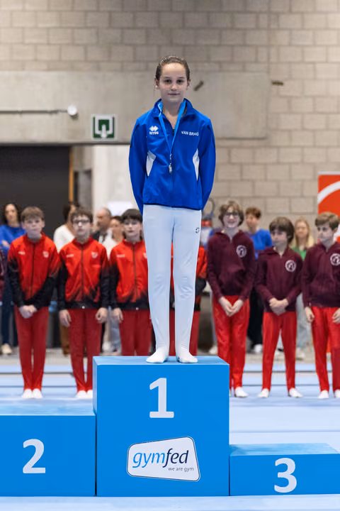 Young gymnast in blue tracksuit stands proudly on first place podium at gymfed event with teammates watching behind