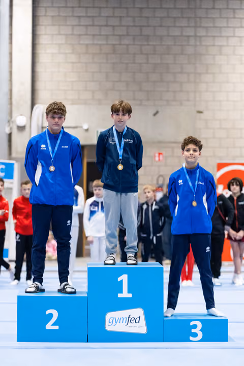 Three young male gymnasts stand on award podium wearing medals, first place in dark tracksuit, others in blue
