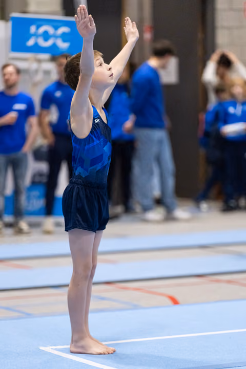 Young gymnast in blue leotard performs an upward salute with arms raised, standing poised on the floor mat.