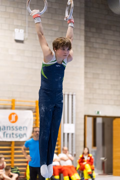 Young gymnast hanging from rings with focused expression, wearing navy uniform with green trim during routine