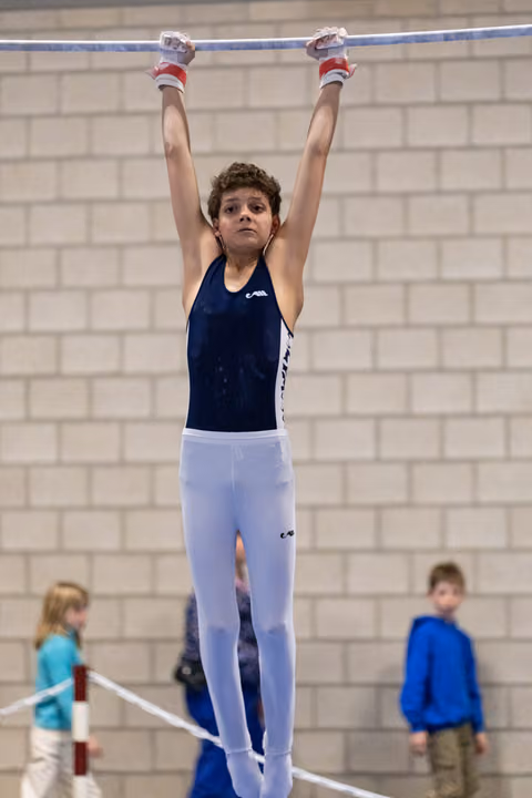 Young gymnast in navy leotard and white pants hangs from high bar with arms extended, focused expression in training gym