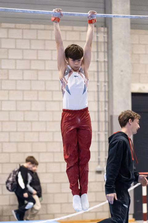 Young gymnast in maroon pants and white leotard hangs from high bar with focused expression during training session