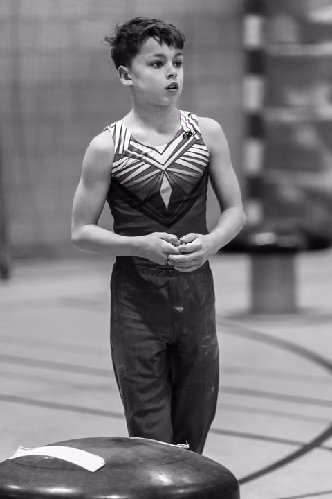 Young gymnast stands composed beside vault, hands clasped, wearing black and white striped leotard in training facility