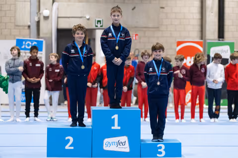 Three young gymnasts stand on victory podium wearing medals and matching navy tracksuits as teammates watch in background