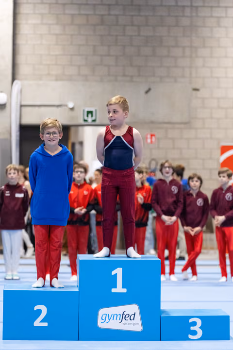 Two young gymnasts stand on a podium in red and blue uniforms, celebrating their achievements with teammates watching behind