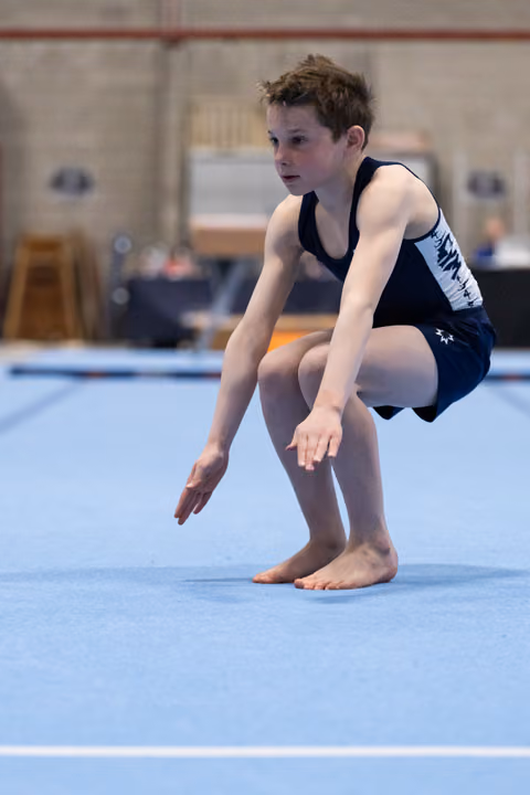 Young gymnast in navy leotard crouches in focused preparation on blue floor mat, arms extended in starting position