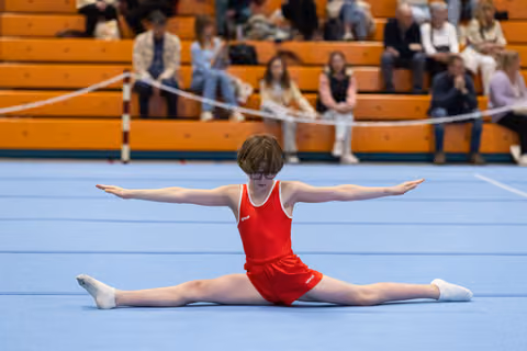 Young gymnast in red leotard performs split on floor mat with arms extended, spectators watching from wooden bleachers