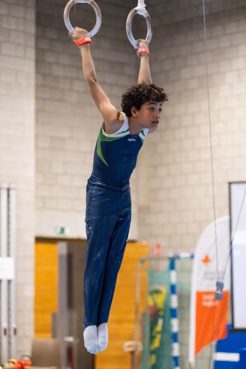 Young male gymnast in navy blue uniform hangs from still rings, demonstrating upper body strength in training facility