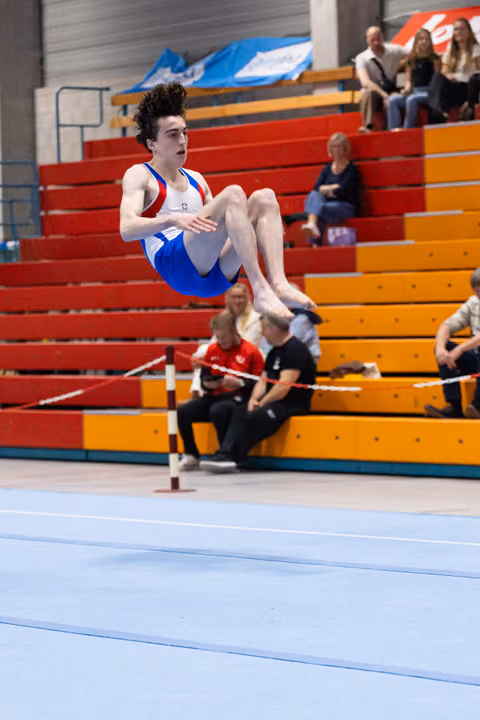 Young male gymnast performs a tucked flip mid-air during floor exercise routine with spectators in bleachers behind