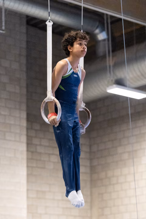 Young gymnast with curly hair holds a static position on the rings, displaying focus and strength during training