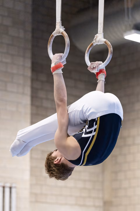 Male gymnast performs inverted tuck position on still rings with extended arms, showing strength and control indoors