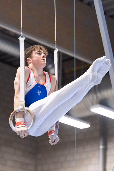 Male gymnast performing rings routine with extended legs, showing intense focus and strength in white pants and red-blue leotard