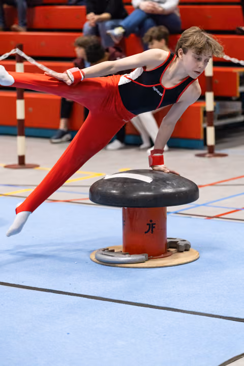 Young gymnast performs a horizontal balance hold on pommel mushroom, demonstrating strength and control in red uniform