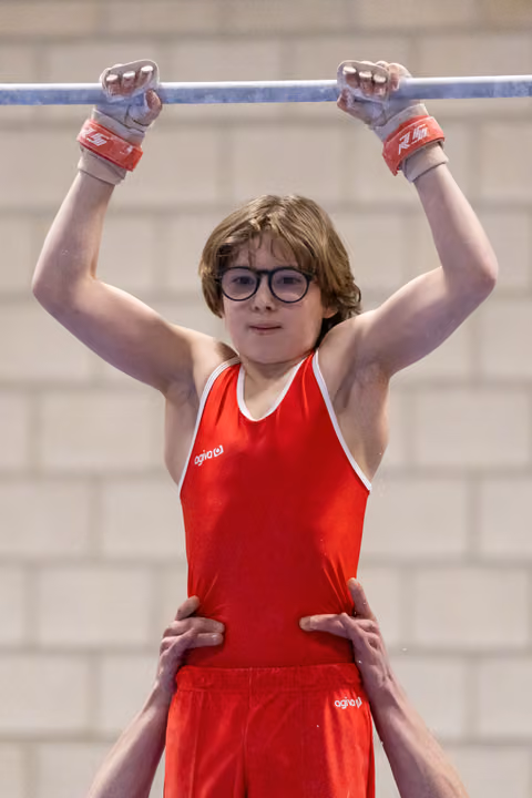 Young gymnast in red leotard and glasses grips horizontal bar while coach supports from below, face showing concentration