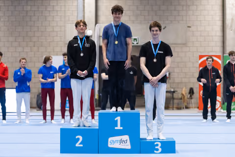 Three young male gymnasts stand on award podium wearing medals, with first place holding gold and teammates in silver and bronze positions