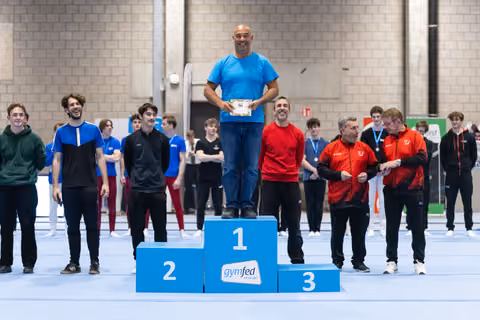 Male gymnast in blue shirt stands atop first-place podium holding trophy, surrounded by competitors in gymnastics hall