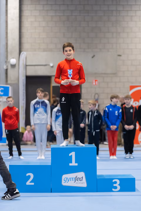 Young male gymnast in red jacket stands on first place podium holding medal, smiling proudly at gymnastics event