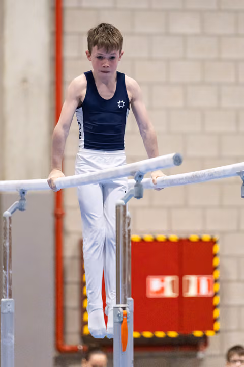 Young male gymnast performs on parallel bars wearing navy and white, displaying intense concentration in indoor gymnasium