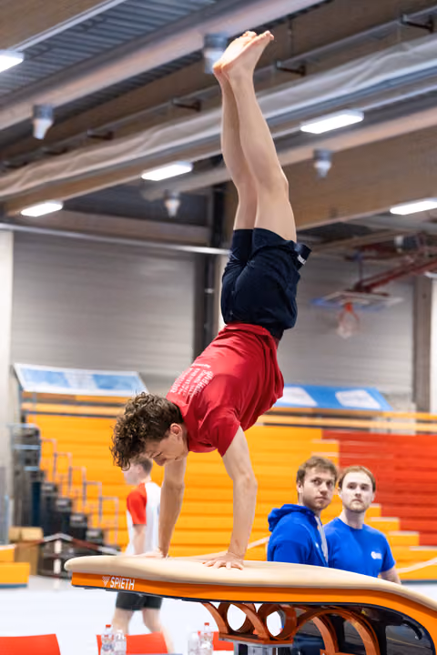 Male gymnast executing a handstand on vault apparatus while two coaches in blue jackets observe from behind
