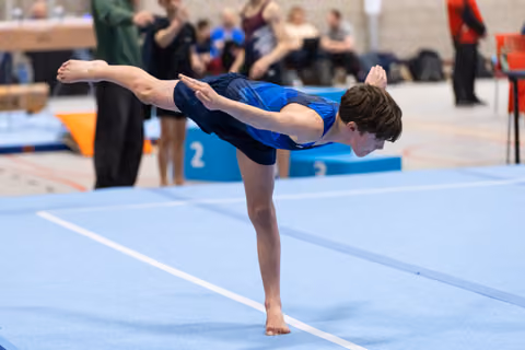 Young gymnast performs a precise arabesque balance on the blue floor mat, demonstrating focus and control during routine