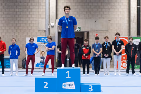 Young gymnast stands proudly on first place podium wearing gold medal, with blue jersey and competitors standing behind