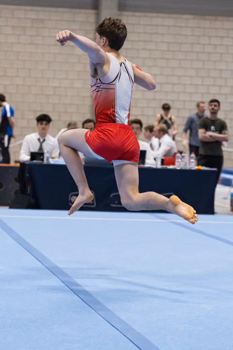 Male gymnast mid-leap during floor routine, arms positioned dynamically, performing before judges and spectators