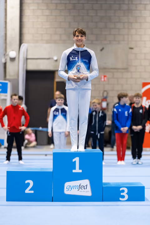 Young gymnast standing proudly on first place podium holding trophy, wearing USA warmup suit at gymfed event