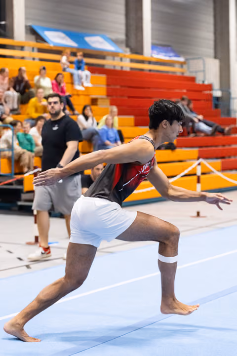 Male gymnast in red and white leotard performs dramatic lunge pose on floor mat, arms extended, with spectators in bleachers