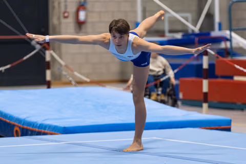 Young gymnast performs an arabesque balance on floor, arms extended gracefully, wearing light blue and navy leotard