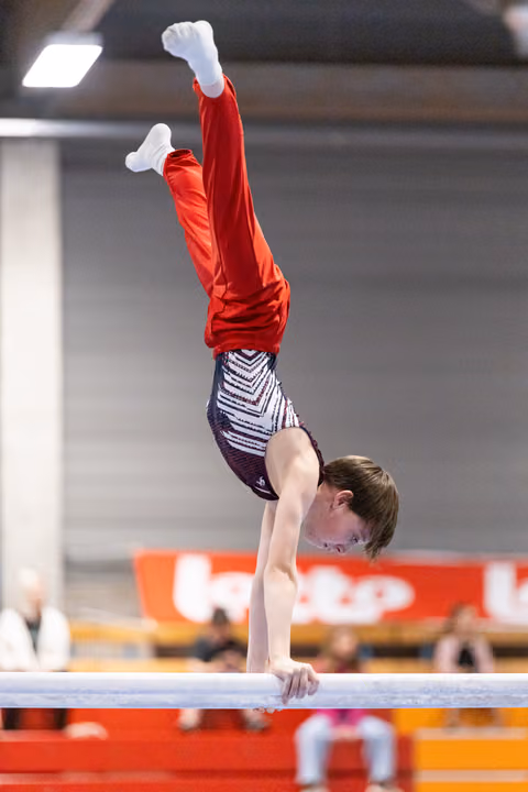 Young gymnast performs handstand on parallel bars wearing red top and patterned shorts in indoor training facility