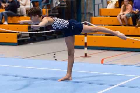 Young gymnast holds a horizontal arabesque balance position with extended arms and leg on the competition floor