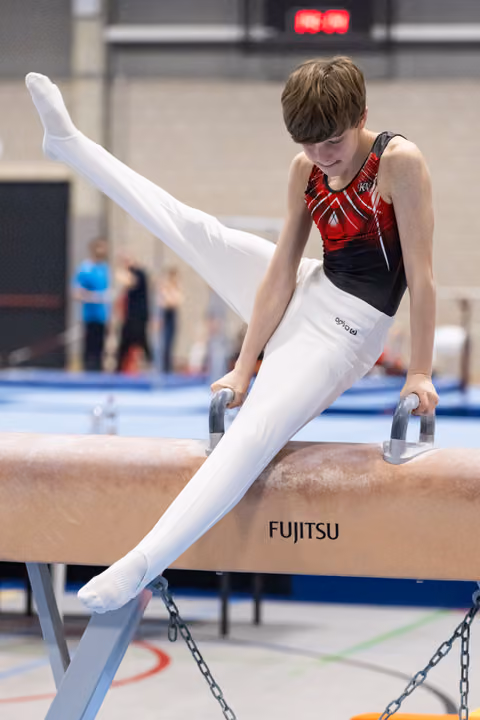 Young male gymnast performing on pommel horse with legs extended upward, wearing red and black leotard in training facility