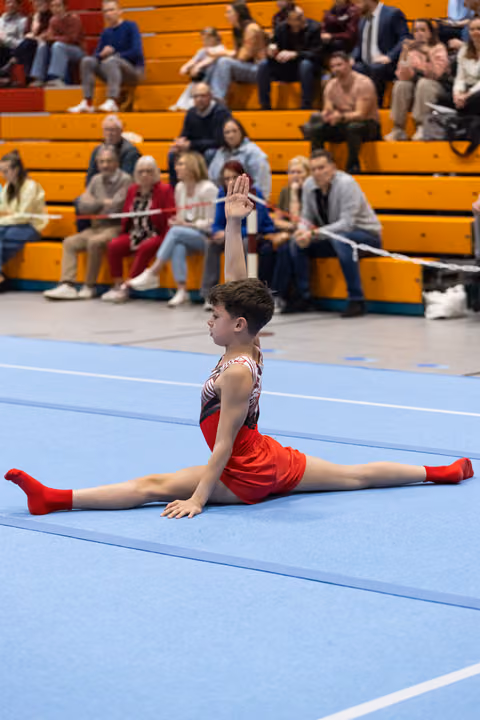 Young gymnast in red performs split on blue floor mat with one arm raised as audience watches from wooden bleachers