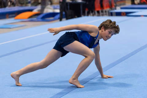 Young gymnast in blue leotard performs a dynamic lunge with extended arms on the spring floor mat