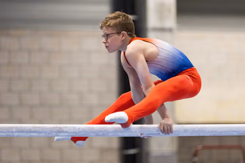 Young male gymnast in orange and blue leotard performs on horizontal bar, body extended horizontally with intense focus