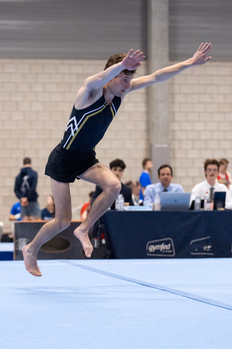 Male gymnast performs powerful leap on floor exercise with arms raised, judges watching from table in background