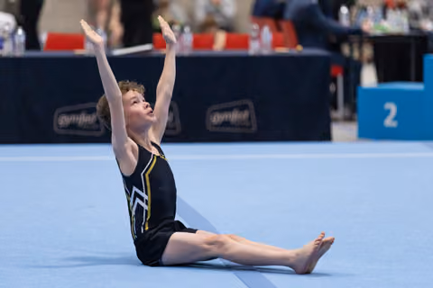 Young gymnast in black leotard performs floor routine, sitting with legs extended and arms raised gracefully overhead